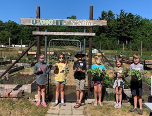 A group of six school-aged children outside in front of a community garden entrance standing and holding leafy greens.